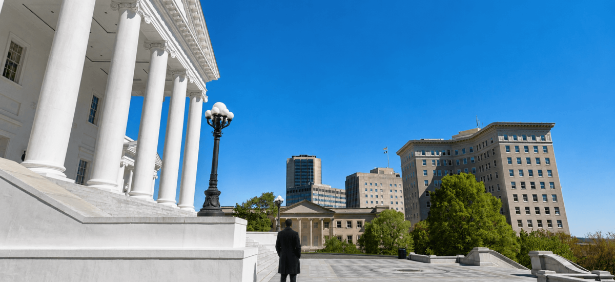 A lone watchman in a dark overcoat stands on the steps of the Virginia State Capitol, looking out over the city of Richmond.